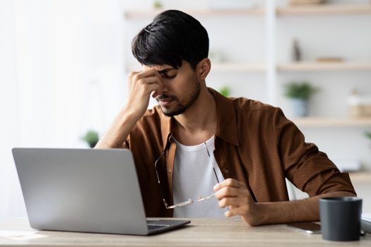 Tired indian man sitting in front of laptop, touching head